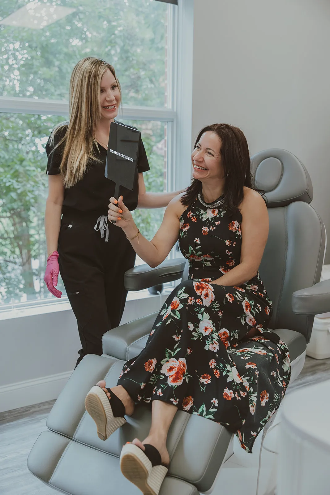 Amy Palacios, FNP, holds up a Mobile Aesthetics hand mirror for a laughing client seated in the treatment chair by the window.